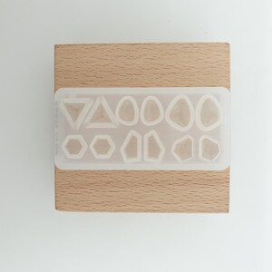 May include: A clear silicone mold with various geometric shapes, including triangles, hexagons, and organic forms. The mold is set on a light brown wooden block, providing a contrasting background. Ideal for resin art and crafting projects.