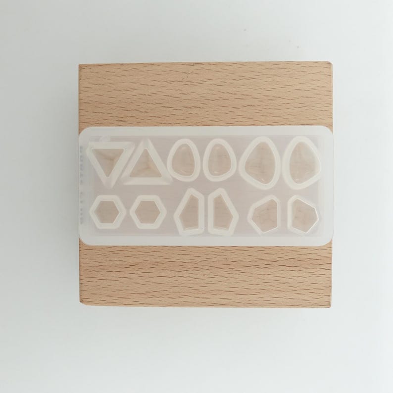 May include: A clear silicone mold with various geometric shapes, including triangles, hexagons, and organic forms. The mold is set on a light brown wooden block, providing a contrasting background. Ideal for resin art and crafting projects.