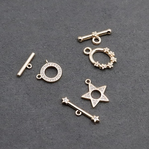 Puede incluir: Una colección de hallazgos de joyería dorados, que incluyen cierres de palanca, un dije en forma de estrella, un anillo floral y un dije circular, todos con pequeños adornos transparentes. Los hallazgos están dispuestos sobre una superficie gris oscuro.