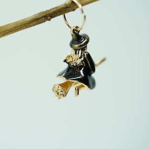 May include: A gold-toned witch earring with a black dress and hat, riding a broom. The earring is suspended from a gold hoop, with the witch's hair and broom details in gold. The earring is a Halloween-themed accessory.