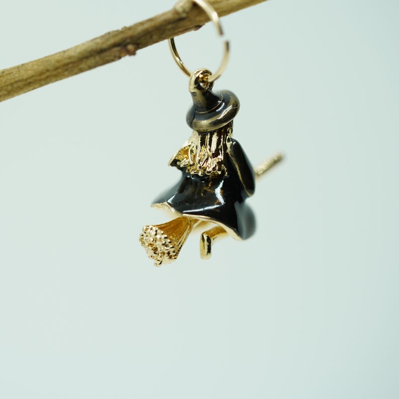 May include: A gold-toned witch earring with a black dress and hat, riding a broom. The earring is suspended from a gold hoop, with the witch's hair and broom details in gold. The earring is a Halloween-themed accessory.