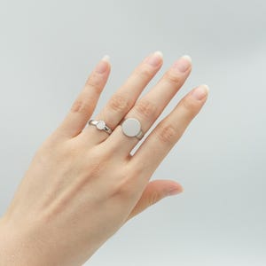 May include: Two silver rings with circular designs. One ring has a small circle, the other a larger, flat circle. The rings are shown on a hand against a plain background. The rings are simple and modern.