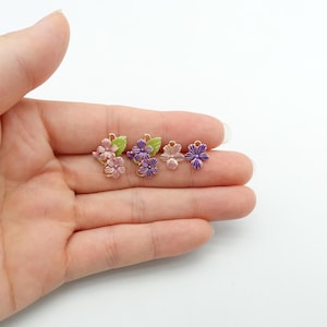 May include: A collection of small, flower-shaped charms in shades of pink, purple, and gold. Each charm features detailed petals and a small loop for attaching to jewelry or other crafts. Some charms include green leaves.