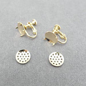 Puede incluir: Componentes de pendientes de clip dorados. Los componentes incluyen un disco plano y redondo con una serie de pequeños agujeros y un mecanismo de clip. Los pendientes están diseñados para orejas no perforadas. Los componentes se muestran sobre una superficie gris.