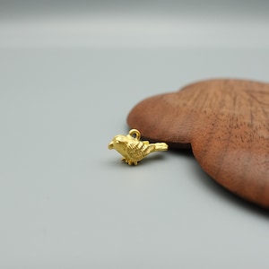 May include: A small, gold-coloured bird charm with a loop for hanging. The charm features detailed, textured feathers and a small beak. The bird is positioned on a grey surface with a wooden object in the background.