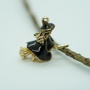 May include: A gold-toned witch charm with black enamel details, riding a broom. The charm features a witch's hat and dress, with a broom made of gold-colored metal. The charm is designed for jewelry making or crafting.