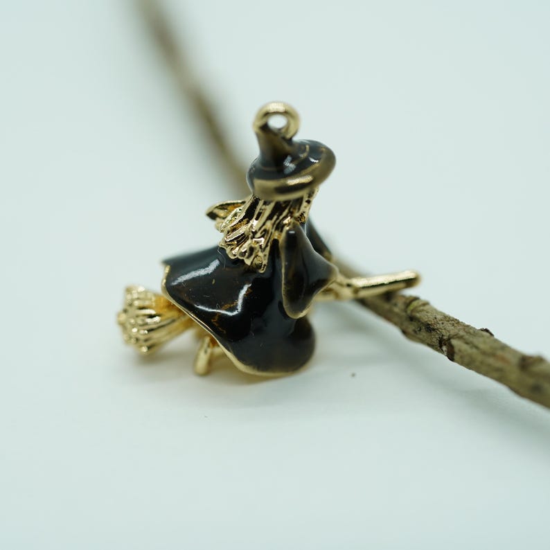 May include: A gold-toned witch charm with black enamel details, riding a broom. The charm features a witch's hat and dress, with a broom made of gold-colored metal. The charm is designed for jewelry making or crafting.