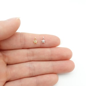 May include: Two small star-shaped charms, one gold and one silver, are shown. Each charm has a small loop at the top for attachment. The charms are displayed against a white background.