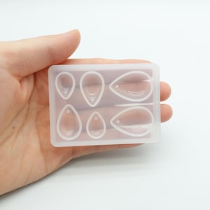 May include: A clear, rectangular silicone mold with six teardrop-shaped cavities. The mold is held in a hand against a white background. The cavities vary in size and shape, designed for crafting earrings or pendants.