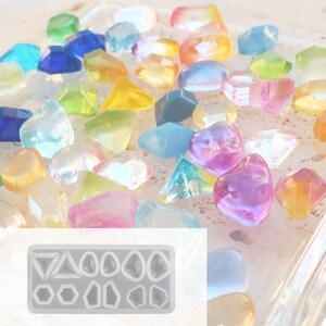 May include: A collection of colorful, translucent gemstone-shaped resin pieces in various shapes and sizes. The gemstones are in shades of blue, green, yellow, orange, pink, and purple. A silicone mold with geometric shapes is in the lower left corner.