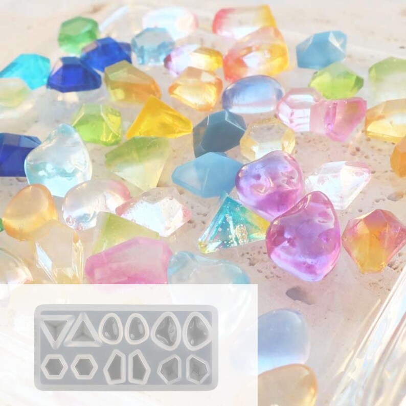 May include: A collection of colorful, translucent gemstone-shaped resin pieces in various shapes and sizes. The gemstones are in shades of blue, green, yellow, orange, pink, and purple. A silicone mold with geometric shapes is in the lower left corner.