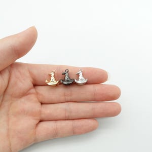Könnte beinhalten: Drei Miniatur-Hexenhut-Anhänger in Gold, Schwarz und Silber. Diese kleinen Anhänger sind mit einer spitzen Krone und einem breiten Rand versehen. Perfekt für Schmuck oder Bastelprojekte zum Thema Halloween. Die Anhänger werden auf hellem Hintergrund gezeigt.