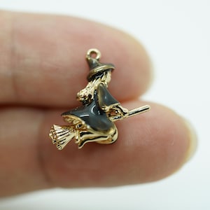 May include: A gold-colored witch charm with black enamel details. The witch is depicted riding a broom, wearing a pointed hat and a flowing dress. The charm is likely for jewelry making or a Halloween-themed craft project.