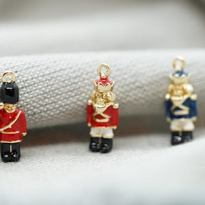 May include: Three gold-colored metal charms shaped like toy soldiers. The soldiers are wearing red, blue, and black uniforms with gold accents. The charms are perfect for adding a festive touch to jewelry or crafts.