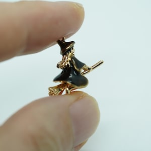 May include: A gold-colored witch charm with a black dress and hat, riding a broom. The charm is small and detailed, with a shiny finish. The witch's hair is gold, and the broom is made of gold-colored metal.