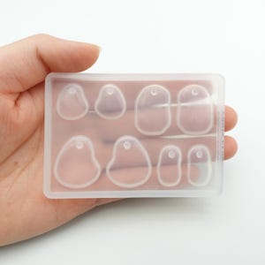 Peut inclure: Un moule en silicone transparent de forme rectangulaire contenant huit cavités en forme de pendentif. Chaque cavité comporte un petit trou en haut pour attacher une chaîne ou un cordon. Le moule est tenu dans une main.
