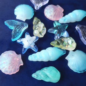 May include: Assortment of translucent seashell and starfish shaped decorative pieces in shades of blue, pink, and yellow. The pieces are scattered on a dark blue surface, creating a coastal theme.