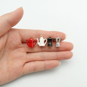 Può includere: Quattro mini ciondoli a forma di caffettiera in rosso, bianco, nero e argento. I ciondoli sono tenuti in una mano su sfondo bianco. Ogni ciondolo ha un piccolo anello nella parte superiore per il fissaggio a gioielli o altri lavori artigianali.