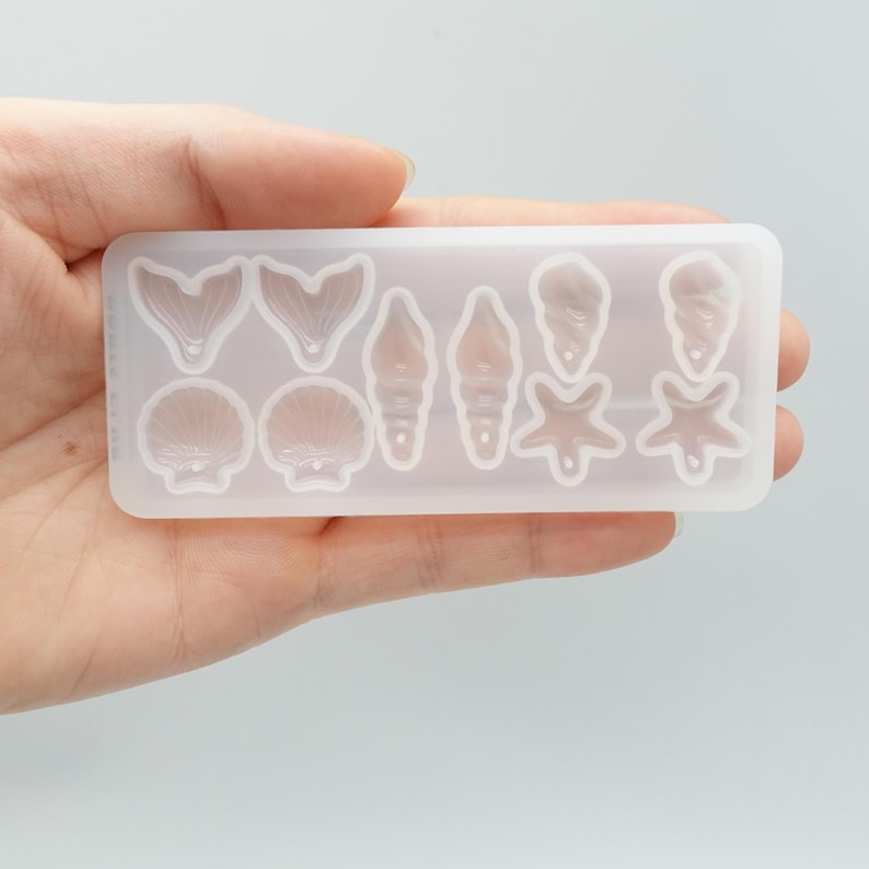 May include: A clear silicone mold with various sea-themed shapes, including mermaid tails, seashells, starfish, and conch shells. The mold is rectangular and designed for crafting resin jewelry or decorative items. The shapes are detailed and ready for casting.