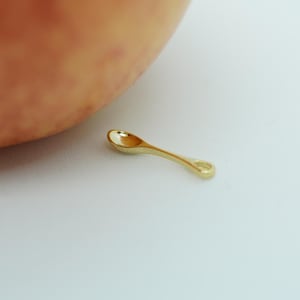 Pode incluir: Uma pequena colher dourada com um cabo curvo e uma tigela arredondada. A colher está colocada sobre uma superfície branca, com uma porção de um pêssego visível no fundo.