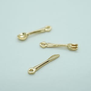 Op de afbeelding: Drie goudkleurige miniatuur bestekbedels: een lepel, een vork en een mes. Elke bedel heeft een kleine lus aan het uiteinde om aan sieraden of andere knutselwerken te bevestigen. De bedels liggen op een lichtblauwe achtergrond.