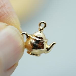 以下が含まれることがあります： 詳細なデザインが施された金色のティーポットチャーム。チャームの上部には、ネックレスやブレスレットに取り付けるためのループが付いています。