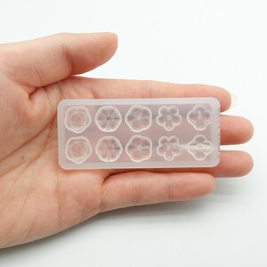 May include: A clear silicone mold with a rectangular shape, featuring nine flower designs. The mold is held in a hand against a white background. The flowers include roses and other floral shapes.