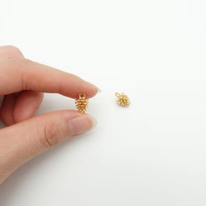 Puede incluir: Dos pequeños colgantes dorados con forma de piña. Uno se sostiene entre los dedos, mientras que el otro descansa sobre una superficie blanca. Los colgantes tienen una superficie texturizada y un pequeño lazo en la parte superior.