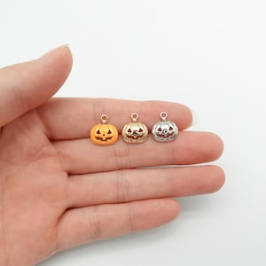 May include: Three miniature pumpkin charms in orange, gold, and silver. Each charm features a carved jack-o'-lantern face and a loop for attachment. Ideal for jewellery making, these charms are displayed against a light background.