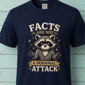 May include: Navy blue t-shirt featuring a raccoon illustration with the text "FACTS ARE NOT A PERSONAL ATTACK" in a vintage style. The raccoon is gray and white with a black mask, and the text is in a serif font.