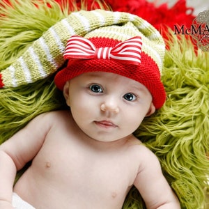 May include: A baby wearing a red and white striped hat with a red bow on top. The hat has a long white tassel. The baby is lying on a green fuzzy surface.