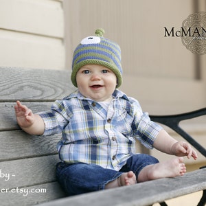 May include: A young child wearing a green and blue knit hat with a white applique on top, sitting on a wooden bench. The child is wearing a plaid shirt and blue jeans. The child is smiling and looking at the camera.