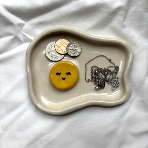 May include: A cream-colored ceramic trinket dish with an irregular shape. The dish contains several coins, a silver necklace, and a yellow, smiling sun-shaped decoration. The dish is on a white surface.