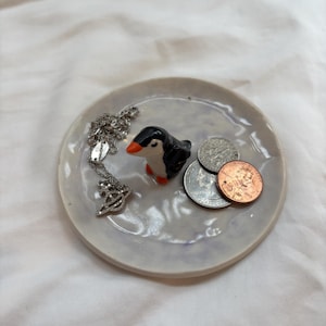 May include: A small, round, ceramic trinket dish with a light purple glaze. A small, black and white penguin figurine with an orange beak sits on the dish, along with a silver necklace and some coins.
