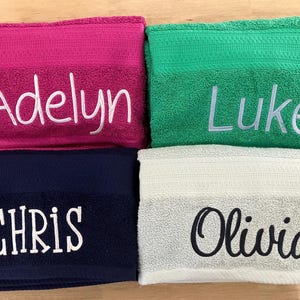 May include: Four personalized bath towels in vibrant colors: fuchsia, green, navy, and off-white. Each towel features an embroidered name in a contrasting color: Adelyn, Luke, Chris, and Olivia.