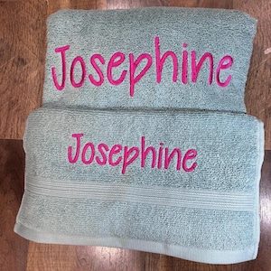 May include: A light green terry cloth towel featuring the name Josephine embroidered in bright pink cursive script. The towel is folded into two sections, with the name Josephine clearly visible on both the top and bottom folded layers.