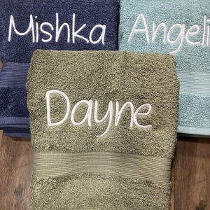 May include: Three folded terry cloth towels in navy blue, light blue, and olive green. Each towel features white embroidered text. The navy towel reads Mishka, the light blue towel reads Angeli, and the olive green towel reads Dayne.