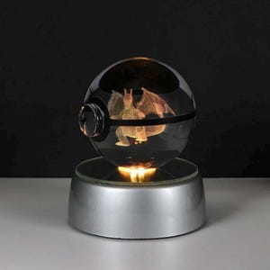 May include: A clear glass sphere with a black band and a 3D image of a dragon inside, resting on a silver base. The base emits a warm, yellow light, illuminating the dragon figure. The sphere is a decorative item.