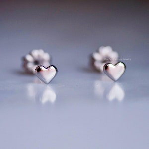 May include: A pair of small, silver heart-shaped stud earrings. The earrings are highly polished and reflect light, sitting on a reflective surface. The earring backs are visible in the background. The earrings are a romantic jewellery item.