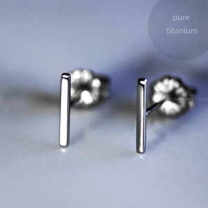 May include: A pair of minimalist silver bar stud earrings. The earrings are made of pure titanium and are displayed on a light gray surface. The text "pure titanium" is in a gray circle in the upper right corner.