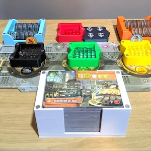 May include: A board game setup with coloured components. Blue, red, green, yellow, and orange trays hold game pieces. A white card holder displays cards with the text "FEDERATION OF ASIA". The game is set on a wooden surface.