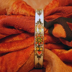 Puede incluir: Una pulsera de brazalete blanca con un diseño floral colorido con corazones, flores y detalles ornamentados. La pulsera se encuentra sobre un fondo de telas naranjas y rojas, creando una estética cálida.