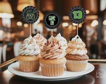 May include: Three cupcakes decorated with different colored frosting and sprinkles. Each cupcake has a round, black and green paper topper with a gaming theme. The toppers say "Level Up", "Game On", and "13".