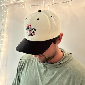 May include: A white and black baseball cap with the text "Ski Mississippi" in red script. The cap features a graphic of a skier and an American flag. The cap has a black brim and a black button on top.