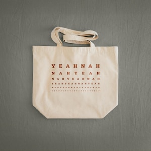 Könnte beinhalten: Cremefarbene Tragetasche mit dem Aufdruck "YEAH NAH" in sich wiederholendem Muster in Dunkelrot. Die Tasche hat zwei Griffe und besteht aus robustem Canvas-Material.