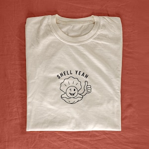 May include: White t-shirt with a black graphic of a smiling clam with a thumbs up and the text "Shell Yeah".