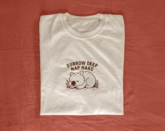 May include: White t-shirt with a brown graphic of a sleeping wombat and the text "Burrow Deep Nap Hard".
