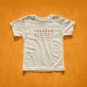 May include: White t-shirt with the words "YEAH NAH" in a repeating pattern in red. The shirt is centered on an orange background. The text is in a sans-serif font.