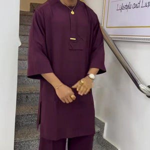 May include: A burgundy two-piece outfit, comprising a tunic and wide-leg trousers, complemented by a matching cap. The outfit has a gold accent on the tunic and is worn with gold-rimmed glasses and a gold necklace. The outfit is part of the "Lifestyle and Luxury" collection.