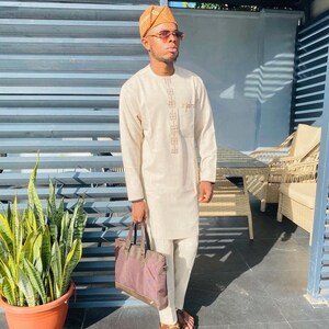 May include: A man wearing a beige long-sleeved shirt with a brown and white geometric pattern, beige trousers, brown sandals, and a brown hat. He is carrying a brown leather tote bag.